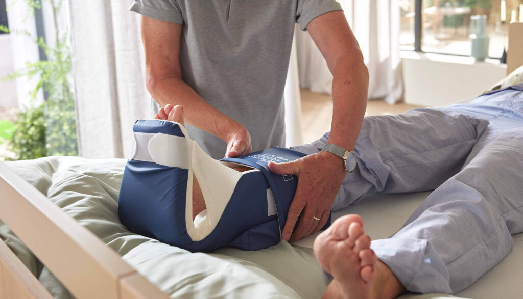 Pfleger befestigt den Fersenfreilagerungsschuh an einer Patientin die im Bett liegt